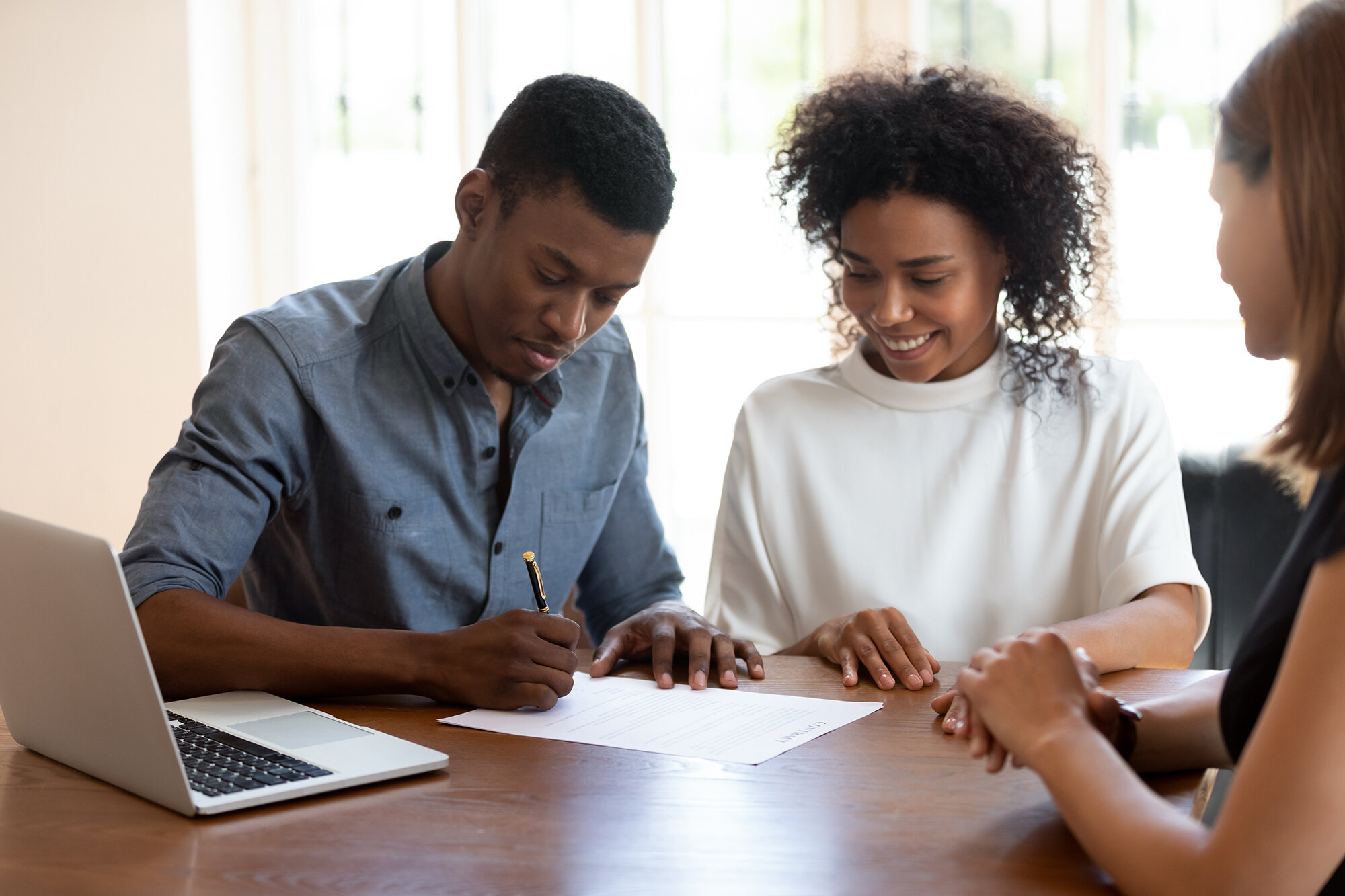 Happy young banking worker, real estate agent or financial advisor watching african ethnicity male client signing agreement. Smiling mixed race woman looking her husband putting signature on contract.